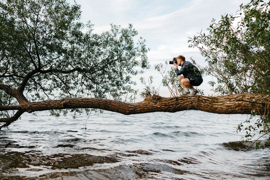 A young man balances on a tree branch taking photos over a scenic body of water.